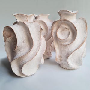May include: Four white ceramic vases with a textured surface and a unique, swirling design. The vases have a ruffled top and a natural, earthy color on the edges, creating a contrast with the white. The vases are arranged closely together.