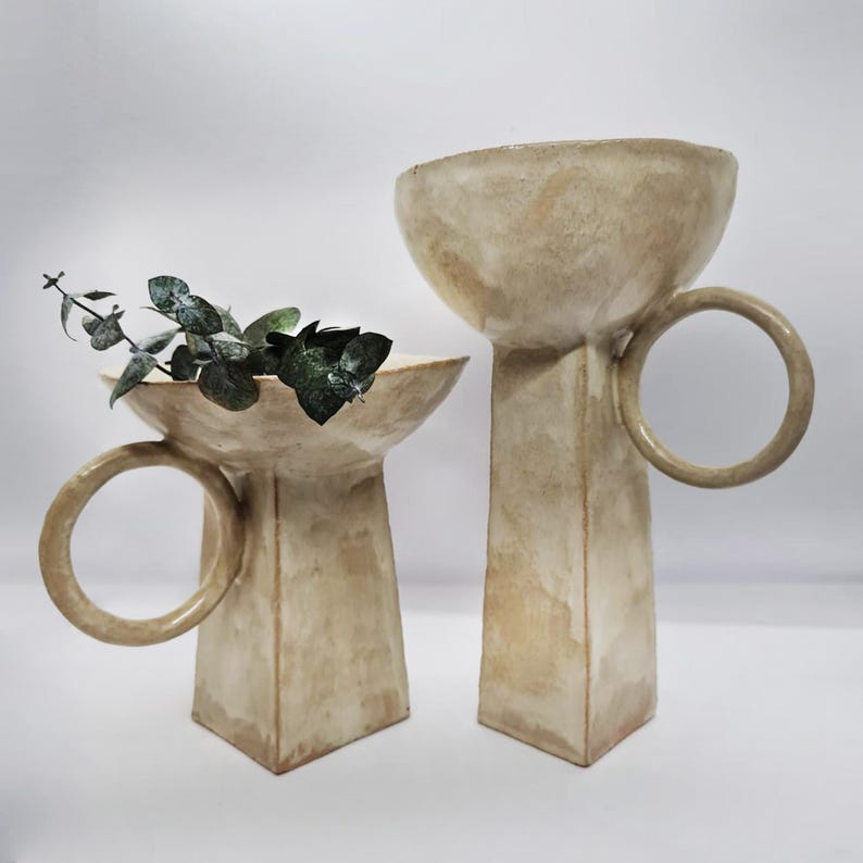 May include: Two beige ceramic vases with a unique design. Each vase has a wide, shallow bowl shape atop a rectangular base. One vase features a ring handle. The vases are displayed on a white surface, and one holds green leafy sprigs, showcasing a minimalist aesthetic.
