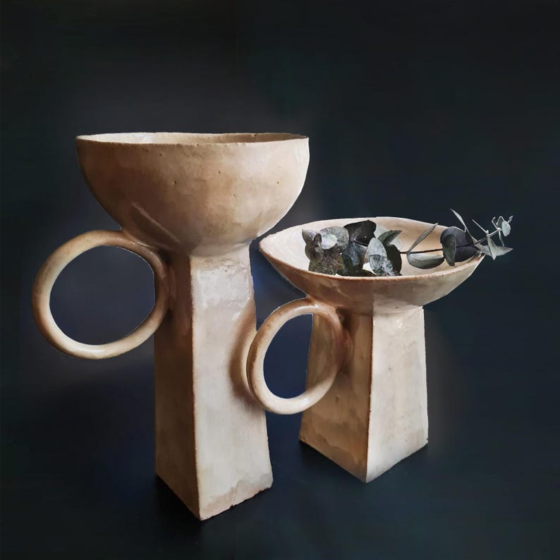 May include: Two beige ceramic pedestal bowls with circular handles. One bowl is empty, and the other contains dark gray stones and a sprig of greenery. The bowls have a textured surface and are set against a dark background.
