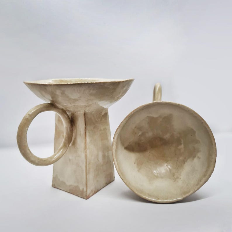 Puede incluir: Dos piezas de cer&aacute;mica hechas a mano. Una es un pedestal beige de base cuadrada con un asa circular y un cuenco ancho y poco profundo. La otra es un cuenco a juego con un asa peque&ntilde;a. Ambos tienen un acabado mate texturizado.