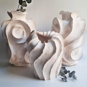 May include: Three off-white ceramic vases with textured, matte surfaces. The two taller vases feature organic, swirling shapes and wide openings, while the shorter, central vase has a distinct spiral, pleated design. Dried eucalyptus stems are visible.