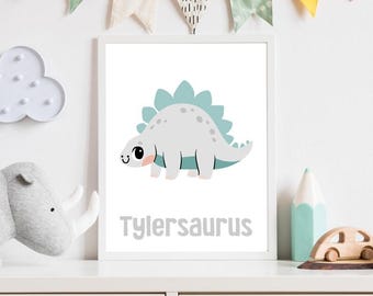 May include: A white framed print with a cute cartoon dinosaur with a teal green back and a light gray body. The dinosaur has big eyes and a pink blush. The text "Tylersaurus" is printed below the dinosaur.