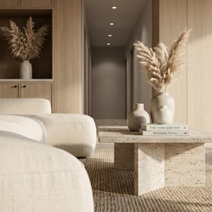 May include: A cream-colored sofa sits beside a square, speckled beige stone coffee table. On the table are two small ceramic vases, a dried pampas grass arrangement, and two stacked books titled The Wabi Sabi Way and Minimalist Forms.