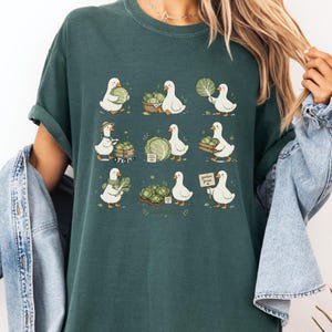 May include: A dark green t-shirt featuring a graphic print of nine white ducks interacting with heads of cabbage in various ways, including holding leaves, pushing a cart, and carrying a sign that reads garden fresh in a cursive script.