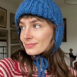 Könnte beinhalten: Eine blaue Strickmütze mit Ohrenklappen und einem Bindeband. Die Mütze ist aus einem groben Strickmaterial gefertigt und hat einen leuchtenden Blauton. Die Mütze ist so konzipiert, dass sie die Ohren bedeckt und unter dem Kinn gebunden wird.
