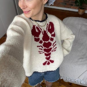 Puede incluir: Un suéter de punto blanco con un diseño de langosta roja. El suéter tiene un corte holgado y un cuello redondo.