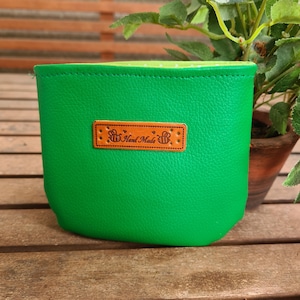 Könnte beinhalten: Ein leuchtend grüner Beutel mit strukturierter Oberfläche und hellgrünem Innenfutter. Mittig auf der Vorderseite befindet sich ein rechteckiges, hellbraunes Lederetikett mit der Aufschrift Hand Made, flankiert von zwei Bienen und Ziernähten.