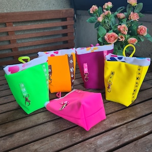Könnte beinhalten: Eine Sammlung farbenfroher Leckerlibeutel für Haustiere in Neongrün, Orange, Magenta, Gelb und Pink. Jeder Beutel hat ein gemustertes Innenfutter und einen passenden Griff. Die Beutel sind mit Pfotenabdruckband und einem Metallverschluss verziert.