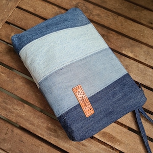 Könnte beinhalten: Eine rechteckige Jeans-Tasche mit Reißverschluss. Die Tasche ist aus verschiedenen Blautönen gefertigt, wodurch ein gestreiftes Muster entsteht. Ein kleines braunes Lederetikett mit dekorativen Herzformen ist vorne angebracht.
