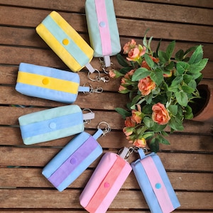 Könnte beinhalten: Eine Sammlung bunter, rechteckiger Täschchen mit Schlüsselanhängern. Die Täschchen sind in verschiedenen Pastellfarben gehalten, darunter Gelb, Blau, Rosa und Lila, mit kontrastierenden Streifen und Knopfverschlüssen. Ein Strauß orangefarbener Rosen steht daneben.