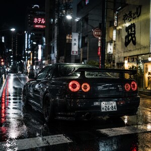 May include: A black sports car with illuminated rear lights parked on a wet street at night. The car's number plate reads "26-84". Buildings with Japanese signage and neon lights line the street.