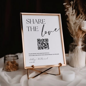 May include: A white sign with a gold frame on a gold easel. The sign says "Share the love" and has a QR code. The text below the QR code says "Please share your photos with us. Scan this code with your smartphone to upload.  Ashley & Nathaniel 01.20.2028"
