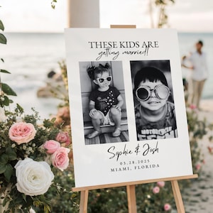 May include: A white wedding sign on a wooden easel, featuring two black and white photos of children wearing sunglasses. The sign reads "THESE KIDS ARE getting married!" and includes the names "Sophie & Josh" and the date and location.