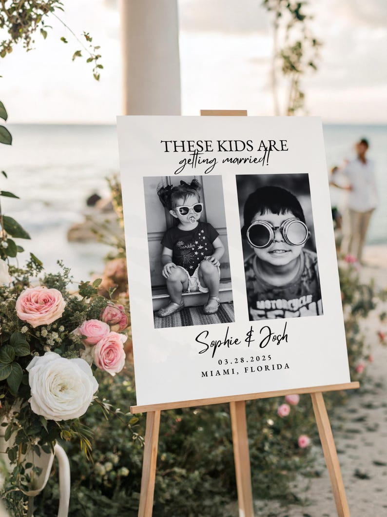 May include: A white wedding sign on a wooden easel, featuring two black and white photos of children wearing sunglasses. The sign reads "THESE KIDS ARE getting married!" and includes the names "Sophie & Josh" and the date and location.