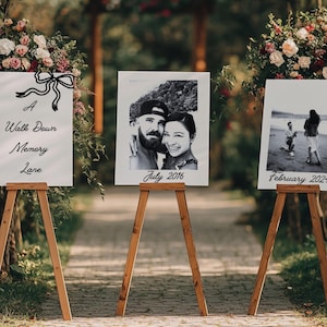 May include: Three wooden easels display framed photos and signs along a stone path. The signs read "A Walk Down Memory Lane" and feature a decorative bow. The photos are dated July 2016 and February 2021, with a couple in each photo.