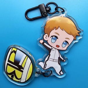 May include: A clear acrylic keychain featuring a cartoon character with blonde hair and blue eyes, wearing a white and black outfit. The keychain also includes a second charm with a yellow and white design and a black clasp.