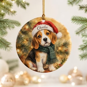 Peut inclure: Une décoration de Noël ronde en céramique illustrant un chiot beagle portant un bonnet de Père Noël rouge avec le nom Charlie inscrit sur le bord blanc et une écharpe en tricot vert, suspendue par un cordon doré.
