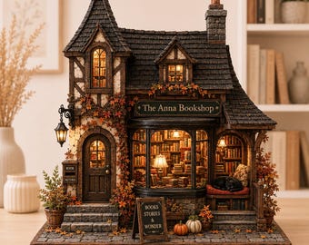 May include: A miniature model of a two-story bookshop titled The Anna Bookshop, featuring a dark shingled roof, stone-textured walls, and warm interior lighting. The exterior is decorated with autumn leaves, a black cat, and a sign reading Books Stories & Wonder.