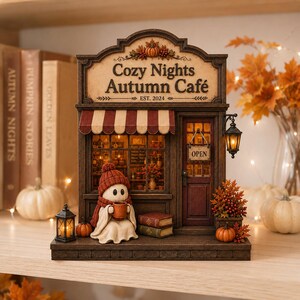May include: A decorative miniature cafe building titled Cozy Nights Autumn Café EST. 2024, featuring a ghost figure wearing a red knit hat and scarf holding a mug, a striped awning, a lantern, stacked books, and an open sign on the door.