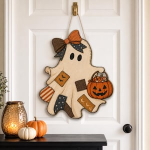 May include: A hanging ghost-shaped wall decoration made of beige fabric with stitched patchwork patterns in orange, brown, and navy blue. The ghost features a large plaid bow, black oval eyes, and carries a jack-o'-lantern bucket filled with candy.