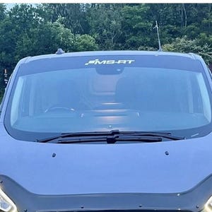 Puede incluir: Una furgoneta gris con el texto "MS-RT" en la parte superior del parabrisas. La furgoneta tiene espejos laterales negros y un protector de capó negro. Los faros están encendidos, estacionada en un estacionamiento.