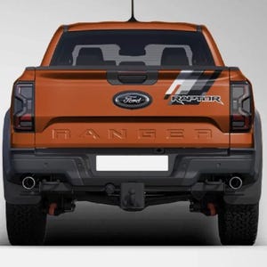 Op de afbeelding: De achterkant van een oranje Ford Ranger Raptor pick-up truck. De achterklep is voorzien van een zwart Ford-embleem, het woord RANGER in reliëf in het midden en een grafische sticker met de tekst RAPTOR. Het voertuig heeft donkere achterlichten, een zwarte bumper, dubbele uitlaatpijpen en een trekhaak.