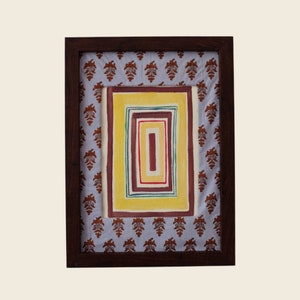 以下が含まれることがあります： 黄色、赤、緑の色合いの同心円状の正方形が並んだ額装されたアート作品。正方形は、茶色の花柄の薄い青色の布で囲まれています。