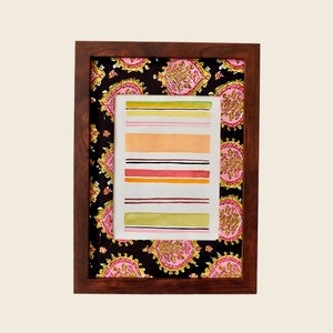 以下が含まれることがあります： 黒い背景にピンクと黄色の花柄が描かれた、額縁に入った水彩画。絵には、カラフルな水平のストライプが描かれている。