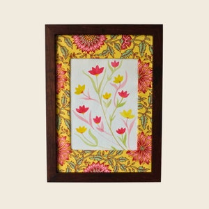 以下が含まれることがあります： 白い背景にピンクと黄色の花が描かれた水彩画の額縁。フレームは濃い木でできており、黄色とピンクの花柄の縁取りが施されています。