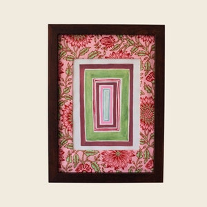 以下が含まれることがあります： ピンクの花柄の縁取りと、緑と茶色の色合いの入れ子になった正方形が並んだ、額装されたアート作品です。