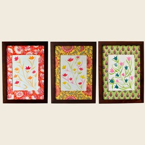 以下が含まれることがあります： 異なる色の花と模様のある縁取りが施された、額装された花のプリントが3つ。フレームはシンプルなデザインの茶色の木です。