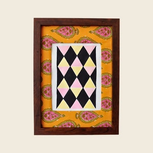 以下が含まれることがあります： 白い背景にピンク、黄色、黒の三角形が幾何学的に並んだフレーム付きの芸術作品。フレームは茶色の木で、マットにはペイズリー柄が施されています。