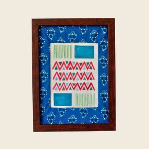 以下が含まれることがあります： 白い背景に赤、ピンク、緑の三角形の幾何学模様が描かれた額縁付きの芸術作品。フレームはダークウッド製で、青い模様のファブリックの縁取りが施されています。