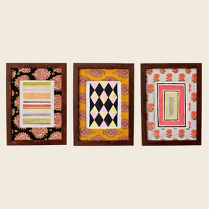 以下が含まれることがあります： 異なる模様の布地の背景を持つ、額装された3枚の絵。最初の絵は白黒の幾何学模様、2枚目はピンクと黄色の幾何学模様、3枚目はピンクと緑の幾何学模様です。