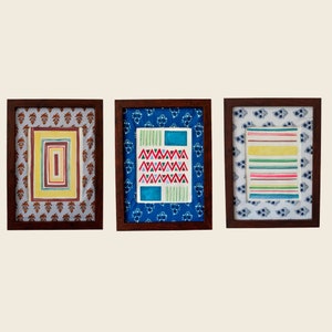 以下が含まれることがあります： 異なるパターンを持つ3つの額縁に入った絵。最初の絵は、黄色の背景に茶色と赤のパターンが描かれている。2番目の絵は、青い背景に白い三角形と線の模様が描かれている。3番目の絵は、白い背景にカラフルなストライプ模様が描かれている。