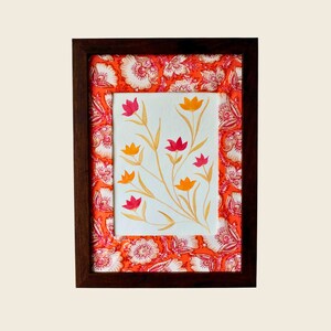 以下が含まれることがあります： 額装された花の芸術作品。この作品は、白い背景に赤とオレンジの色合いで様式化された花の水彩画を特徴としています。フレームは濃い茶色で、マットは花柄の鮮やかなオレンジ色の生地です。