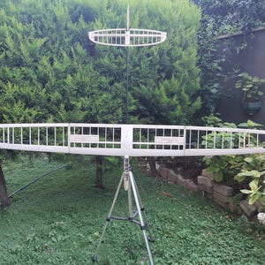 May include: A white model glider with a long, rectangular wing and a smaller wing above. The model is supported by a tripod in a garden setting, with green foliage in the background. The model is designed for flight.