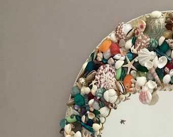 May include: A round mirror with a decorative frame made of seashells, beads, and other ocean-themed trinkets. The mirror is surrounded by a double strand of beaded chains.