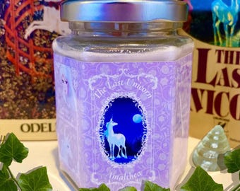 Puede incluir: Un tarro de cristal con tapa plateada y etiqueta lavanda con una ilustración de unicornio. La etiqueta dice "The Last Unicorn" y "Amalthea". El tarro está rodeado de vegetación y una concha.