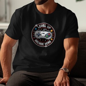 May include: Black t-shirt with a circular graphic featuring a pixelated game controller. The design includes the words "LEVEL UP" and "ARCADE HERO" in a retro style, with stars and a gold border.