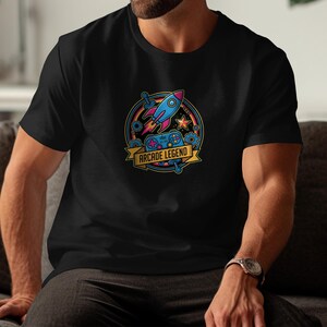 May include: Black t-shirt featuring a colorful graphic with a rocket, game controller, and the text "Arcade Legend." The design includes stars, gears, and a banner. The shirt is short-sleeved.