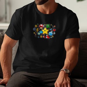 Puede incluir: Camiseta negra con un diseño pixelado colorido de iconos de videojuegos, incluyendo una estrella, una seta, un bloque de interrogación y un rayo. El diseño está centrado en el pecho. La camiseta es de material suave.