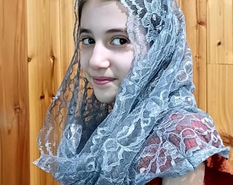 May include: A young person wearing a blue lace head covering and a rust-colored dress. The head covering is draped over the head and shoulders, with intricate floral patterns. The dress has a textured pattern.