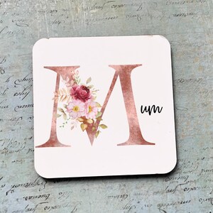 May include: A white coaster featuring a large, rose gold letter "M" adorned with pink and white floral details. The word "um" is written in black script beneath the letter.