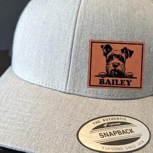 May include: A gray and black baseball cap with a brown leather patch featuring a black and white dog silhouette and the name "BAILEY". The cap has a black mesh back and a white "THE AUTHENTIC YUPOONG SNAPBACK YUPOONG SINCE 1974" label.