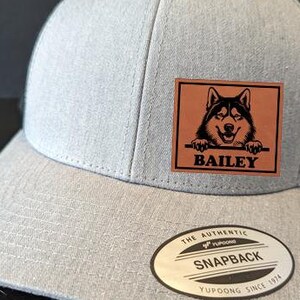 May include: Gray and black snapback hat with a curved brim. The front panel is gray with a brown leather patch featuring a smiling husky dog and the name "BAILEY". The hat has a black mesh back and a "SNAPBACK" label.