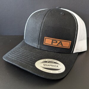 May include: Black and white trucker hat with a brown leather patch that says "PA" on it. The hat has a black brim and a white mesh back. The hat has a Yupoong label that says "The Authentic Yupoong Snapback Yupoong Since 1974".