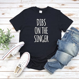 May include: Black t-shirt with white text that reads "Dibs on the singer". The shirt is laid flat on a woodgrain surface with a pair of white trainers and blue jeans.