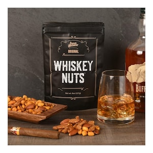 May include: A black bag of "Whiskey Nuts" sits next to a glass of amber liquid and a bottle of whiskey. A wooden tray and a cigar are also on the wooden surface. The bag reads "Swag Brewery Original".