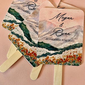 May include: Wedding fans with a watercolor mountain landscape design. The fans are printed with the names Megan & Ryan and the date September 8th, 2025.
